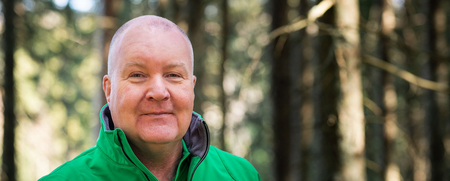 Person i grön jacka står i en solbelyst skog med träd i bakgrunden.