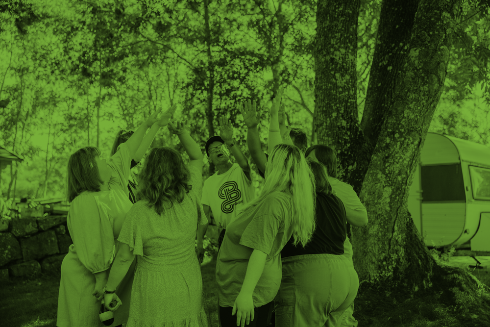 En grupp människor står i en ring utomhus och sträcker upp händerna mot mitten under träd.
