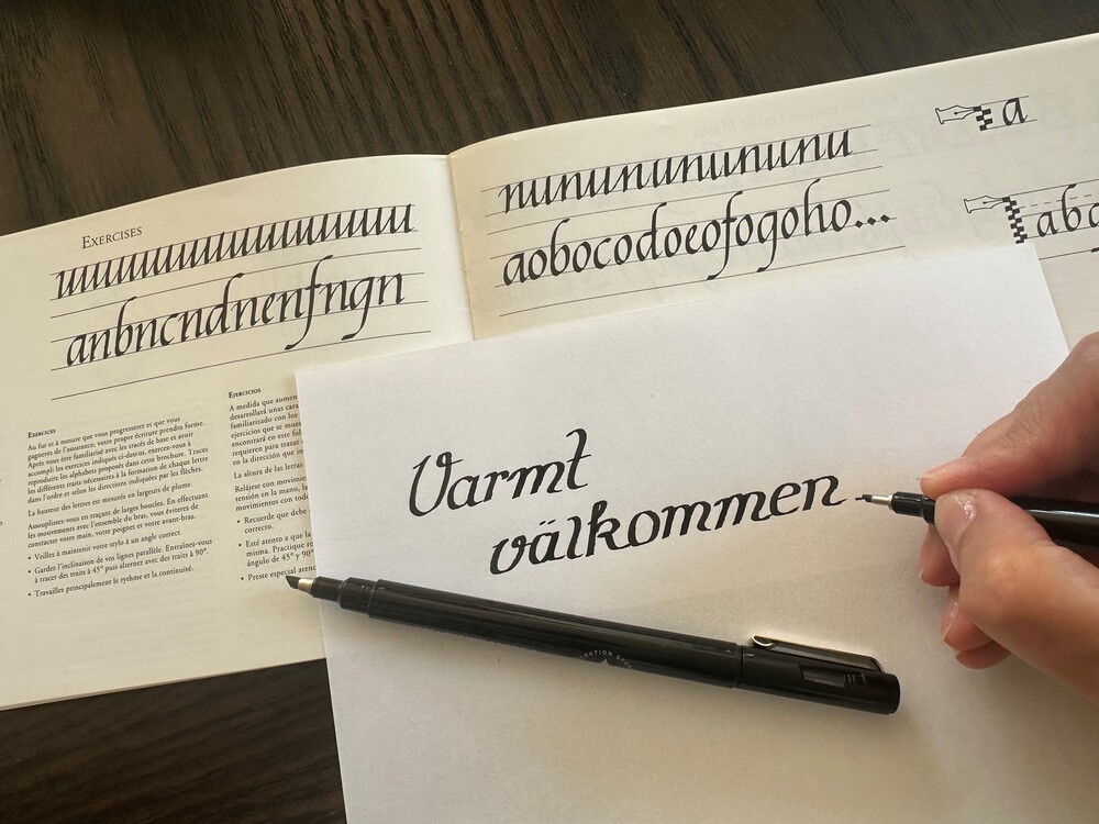 En hand skriver kalligrafi med texten 'Varmt välkommen' på ett papper, med en öppen bok i bakgrunden.