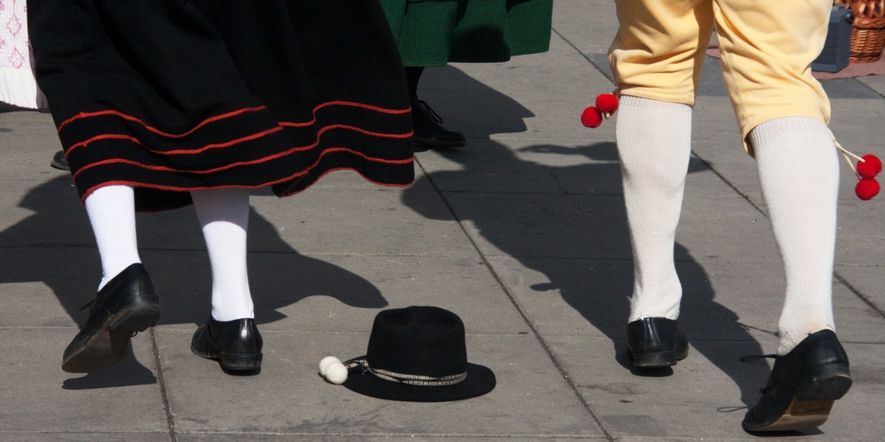 Två personer i traditionella folkdräkter dansar på en stenlagd yta, en svart hatt ligger på marken.