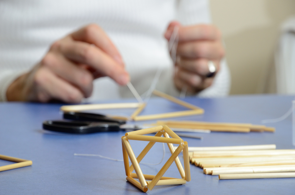 En person bygger en geometrisk form av sugrör och tråd på ett bord, med sax och fler sugrör bredvid.