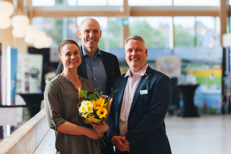 Tre personer står tillsammans inomhus, en kvinna håller en bukett blommor. Ansikten är suddade.