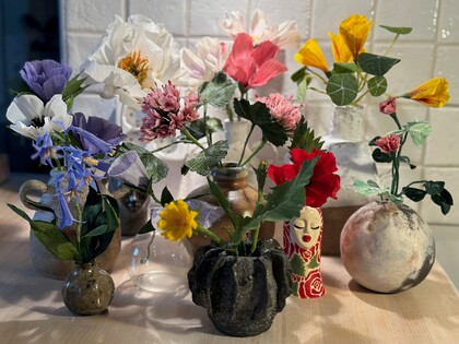 Flera färgglada konstgjorda blommor i olika vaser på ett bord, med en liten figur i keramik.