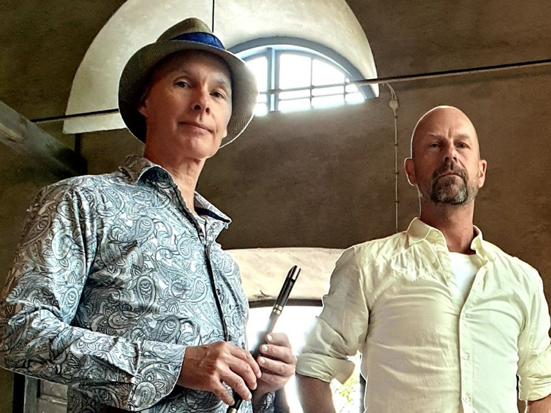 Två män står inomhus framför ett stort fönster, en med hatt och flöjt, den andra i vit skjorta.