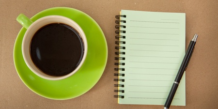 En kopp svart kaffe på ett grönt fat bredvid ett tomt spiralblock och en penna på ett brunt bord.