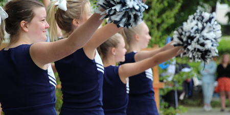 Fyra unga cheerleaders i blå uniformer håller pom-poms och gör en koreograferad rörelse utomhus.