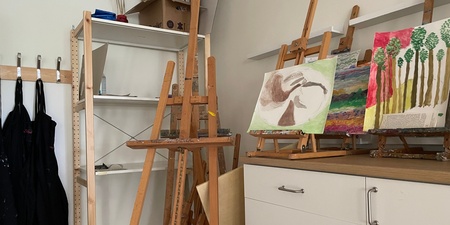 Ett konstnärsrum med stafflier, målningar, svarta förkläden och hyllor.