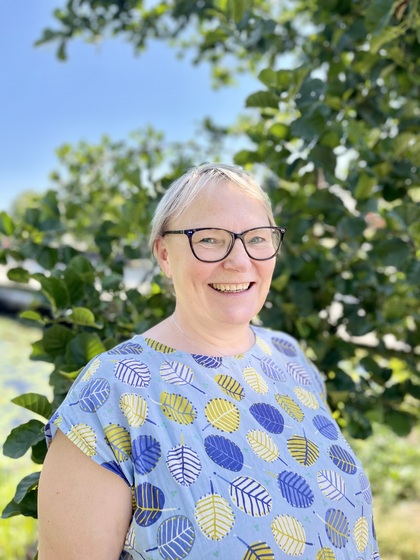 Leende kvinna med glasögon står utomhus framför gröna träd i solsken.