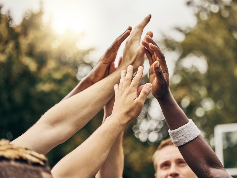 Flera händer från olika personer möts i en high five utomhus, med träd i bakgrunden.
