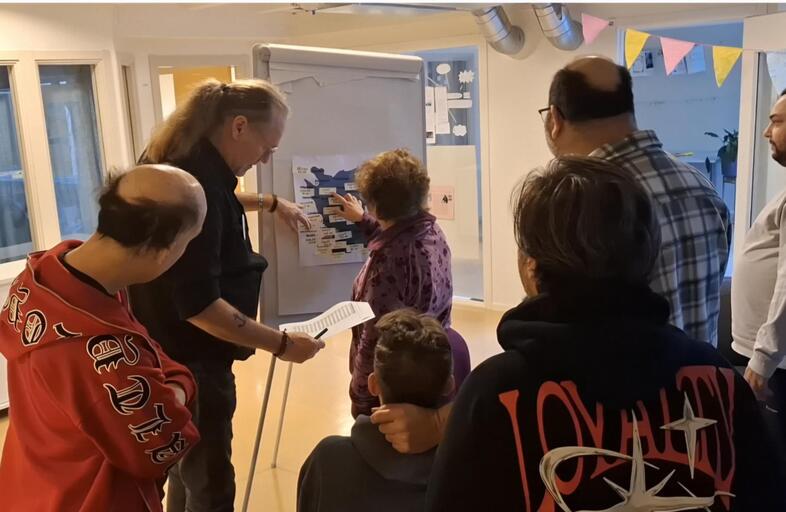 En grupp människor samlas runt en whiteboard i ett ljust rum för en gemensam diskussion.