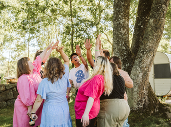En grupp människor står i en ring utomhus och sträcker upp händerna mot mitten under ett träd.