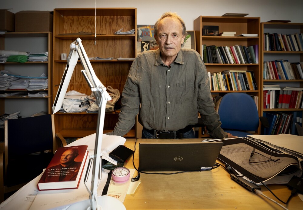 Person står vid ett skrivbord i ett kontor med böcker, papper och en laptop.