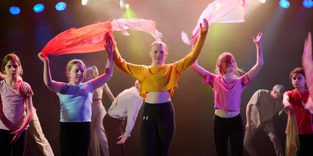 En grupp unga personer dansar på scen med färgglada sjalar under scenbelysning.