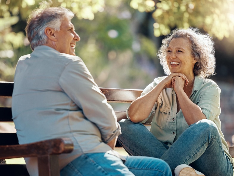 Två äldre personer sitter på en parkbänk och samtalar glatt i solskenet.
