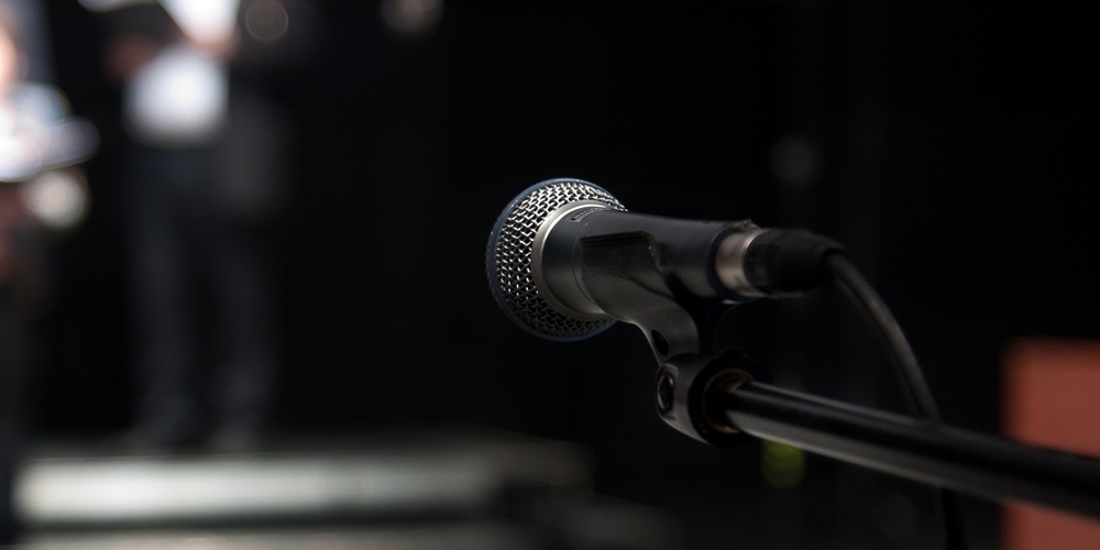 En mikrofon i fokus på en scen med suddiga personer i bakgrunden, troligen en kör eller grupp som förbereder sig.