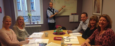 Sex personer sitter runt ett bord med papper och pennor, en person står vid en whiteboard och presenterar.