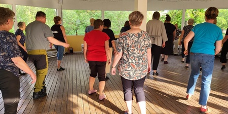 En grupp vuxna dansar tillsammans på ett trägolv i en ljus paviljong med stora fönster.