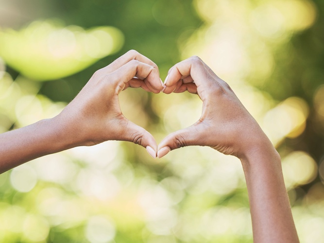 Två händer formar ett hjärta mot en suddig, grön bakgrund utomhus.