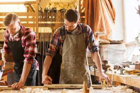 Två personer arbetar med trä i en snickeriverkstad. Båda har förkläden på sig.
