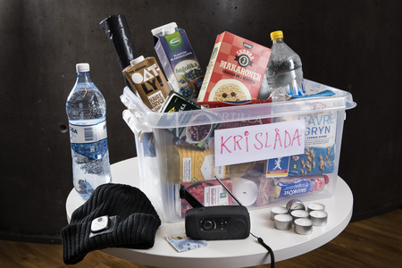 En plastlåda märkt 'KRISLÅDA' fylld med mat, vattenflaskor och nödvändiga saker står på ett runt bord.