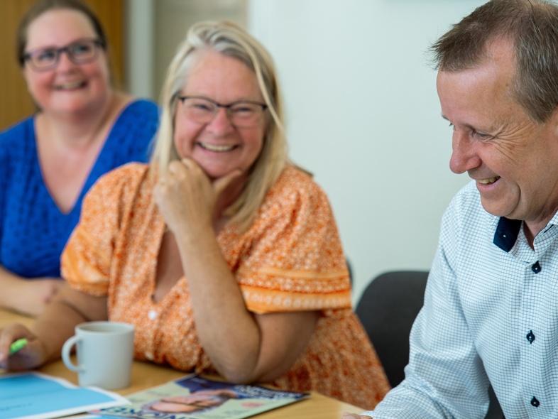 Tre glada vuxna sitter vid ett bord och diskuterar, med papper och pennor framför sig.