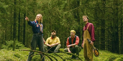 Fyra personer vistas i en grön skog, två sitter på marken och två står, en håller ett musikinstrument.