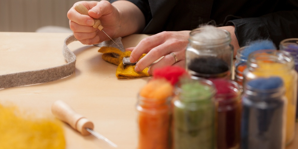 En person arbetar med nåltovning vid ett bord, omgiven av burkar med färgad ull.