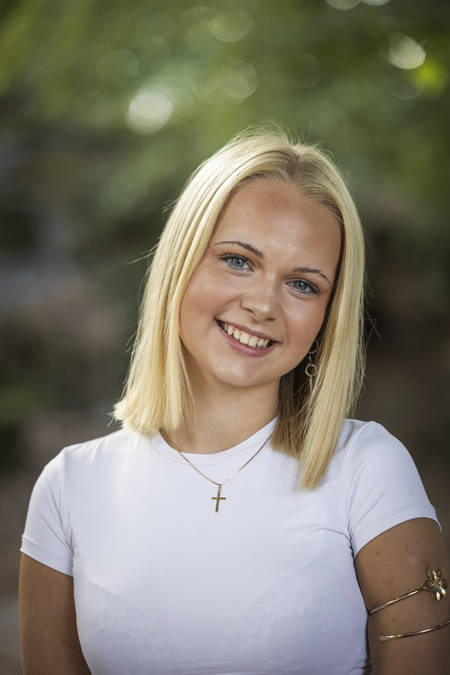 Ung kvinna med blont hår och vit tröja ler mot kameran utomhus.