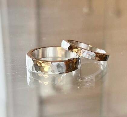 Två ringar i silver och guld med hamrad yta ligger på en reflekterande yta.