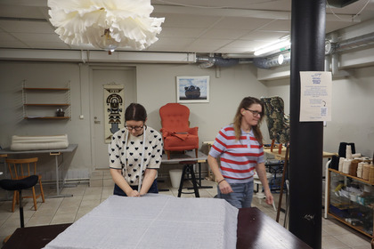 Två personer arbetar med tyg i en ljus syateljé med möbler och verktyg.
