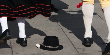 Två personer i traditionella folkdräkter dansar på en stenlagd yta, en svart hatt ligger på marken.