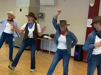 Fem personer dansar linedance inomhus, klädda i cowboyhattar, jeans och scarfar.