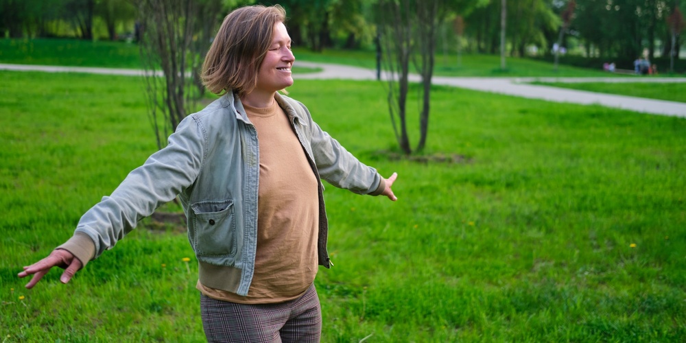 Person står med utsträckta armar i en grön park, ser glad ut och njuter av naturen.
