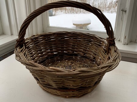 En tom flätad korg med handtag står på ett vitt bord framför ett fönster med snöig utsikt.