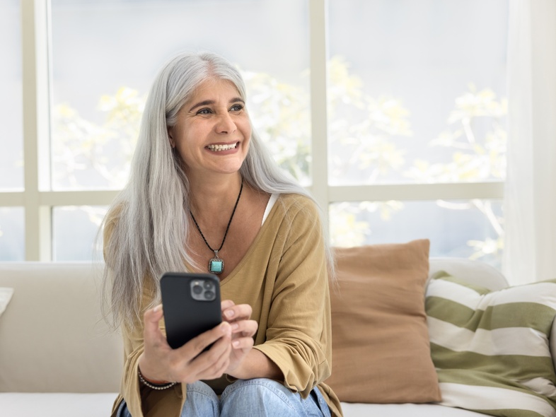Kvinna med långt grått hår sitter i soffa och håller en mobiltelefon i vardagsrum med stora fönster.
