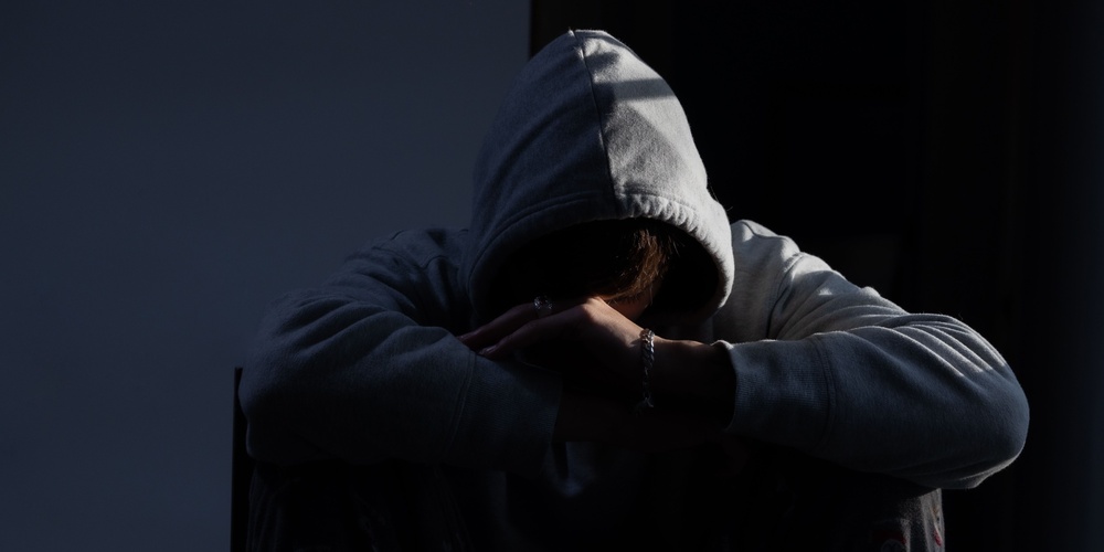 Person i grå hoodie sitter med huvudet nedböjt och armarna korsade, i ett mörkt rum.