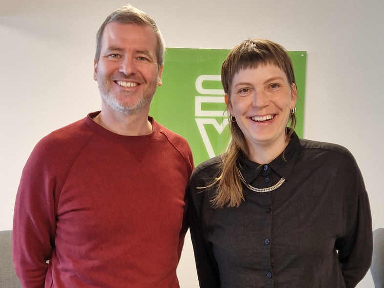 Två personer står bredvid varandra och ler mot kameran i ett ljust rum med grön skylt i bakgrunden.