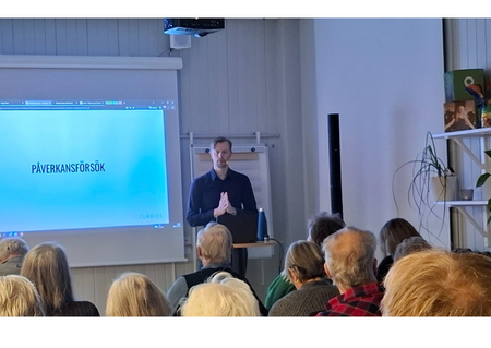 Föreläsning med publik i ett ljust rum, presentation visas på projektorduk.