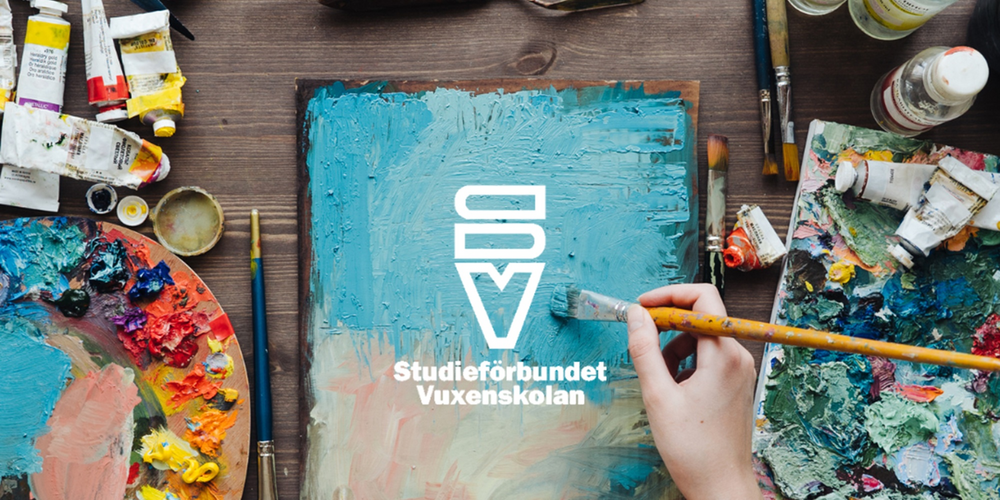 En person målar på en duk omgiven av färgtuber, penslar och konstmaterial på ett bord.