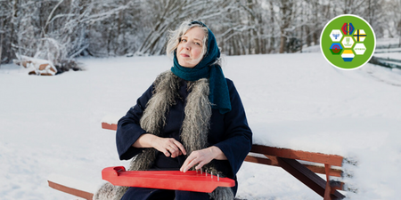 Kvinna i vinterkläder sitter på bänk i snölandskap och spelar ett rött stränginstrument.