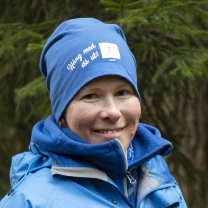 Person i blå jacka och mössa står utomhus i skogsmiljö. 