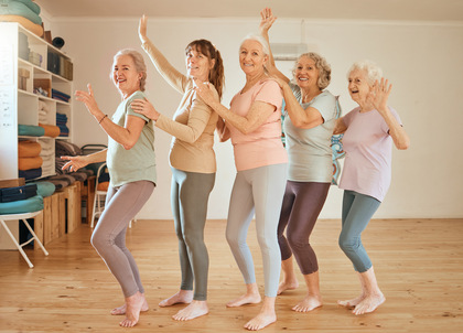 Fem äldre kvinnor gör yoga eller dansar tillsammans i ett ljust träningsrum med trägolv.
