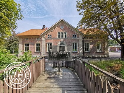 Gammalt tegelhus med spetsigt tak, sett från en träbro omgiven av träd och grönska.