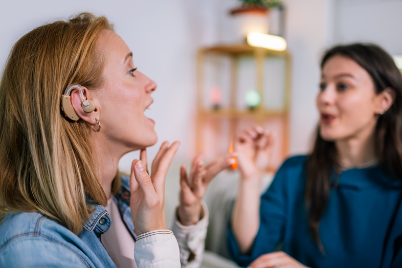 Två personer kommunicerar med teckenspråk, en av dem bär hörapparat, inomhusmiljö.
