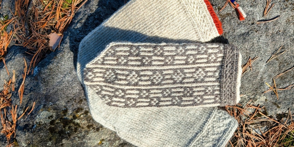 Två stickade vantar i grått och vitt ligger på en sten med tallbarr.