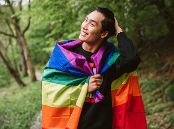 En leende person står utomhus med en regnbågsflagga runt axlarna, omgiven av grönskande träd.
