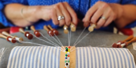 Närbild av händer som tillverkar spets med hjälp av knyppelpinnar och en kudde.