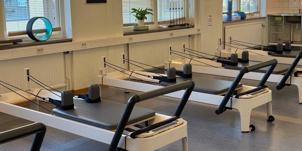 Ett ljust pilatesrum med flera reformermaskiner, stora fönster och träningsredskap på fönsterbrädorna.