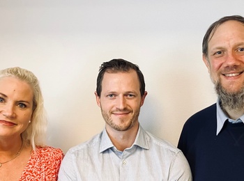 Tre personer står bredvid varandra mot en ljus vägg i ett kontorsliknande rum.