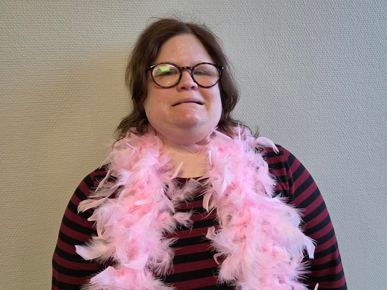 Person med randig tröja och rosa fjäderboa står mot en ljus vägg. Ansiktet är suddat.